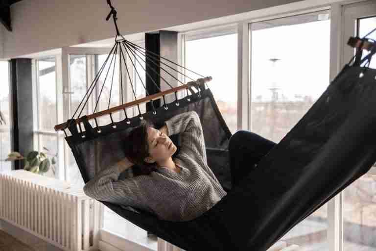 Young lady in casual wear lying in comfortable hammock leaned on hands near window and radiator in apartment in afternoon