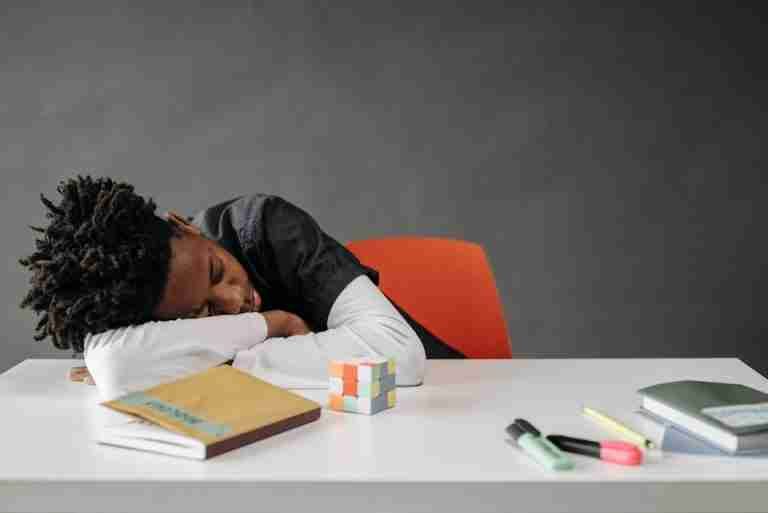 A young boy sleeping at a desk with books and a Rubik's cube, looking weary.