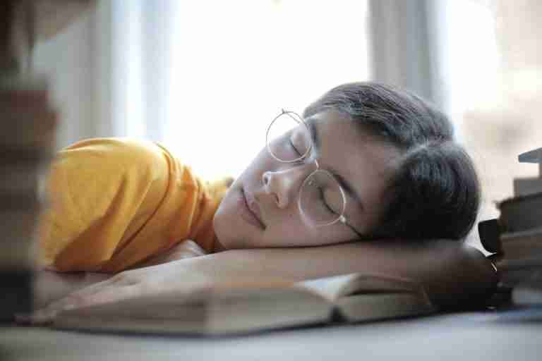 A young woman wearing glasses sleeps on a desk with open books, illustrating fatigue or study break.