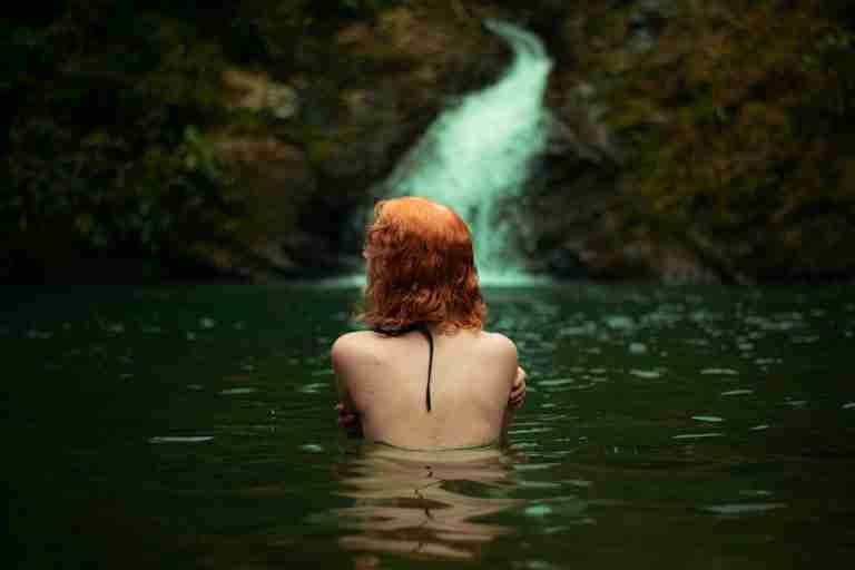 A woman with red hair swims in a tranquil pool at the base of a waterfall surrounded by lush greenery.