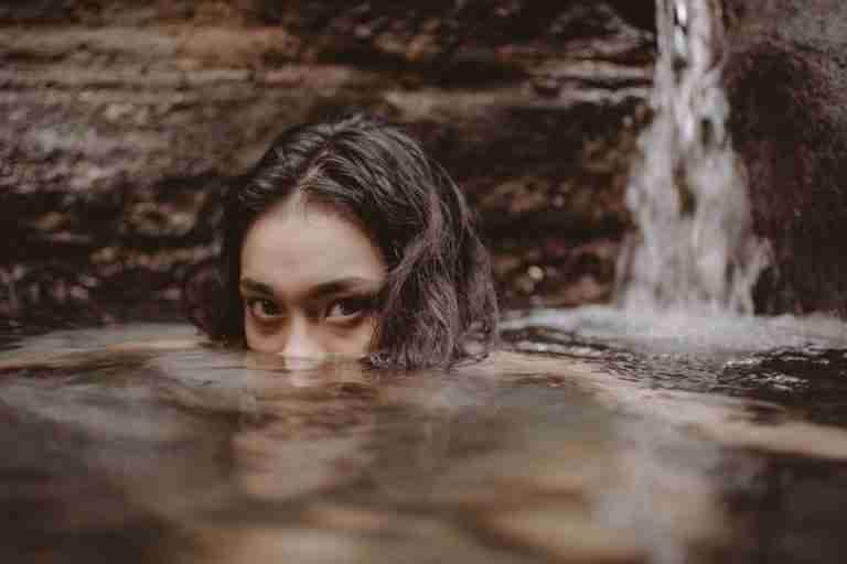 A woman partially submerged in a calm pool, near a waterfall, conveying tranquility.
