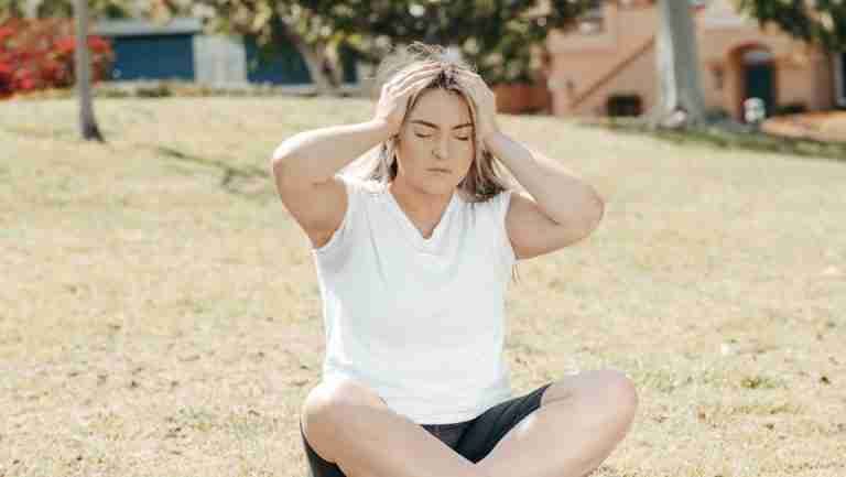 Young woman experiencing a headache while sitting on grass in a sunny park setting.