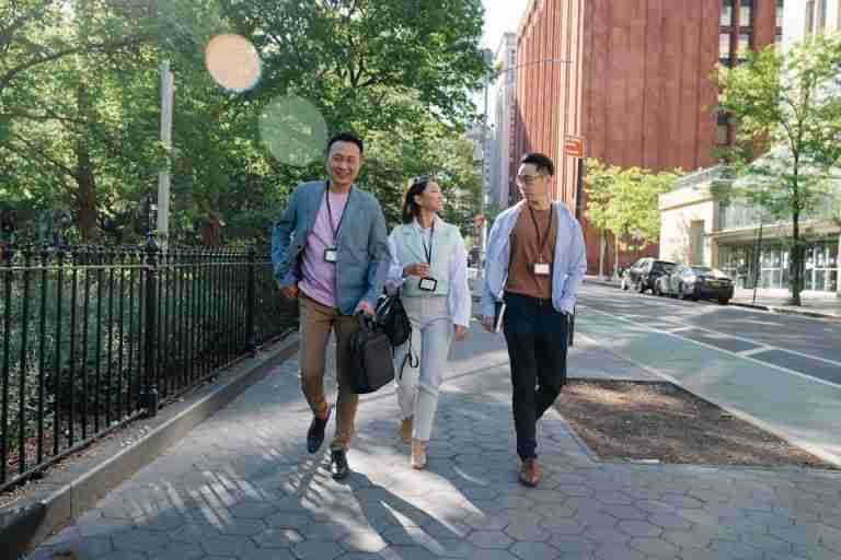 Three adults walking on a city sidewalk during a sunny day break, wearing business attire and ID badges.