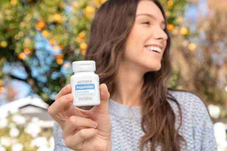 Cheerful woman outdoors displaying a bottle of dōTERRA peppermint dietary supplement.