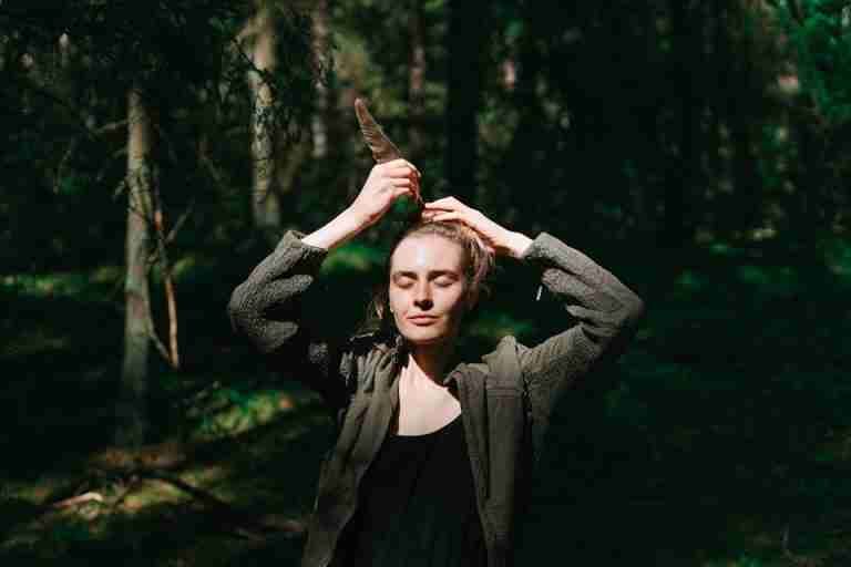 A woman enjoys the sunlight in a serene forest while holding a feather, eyes closed.