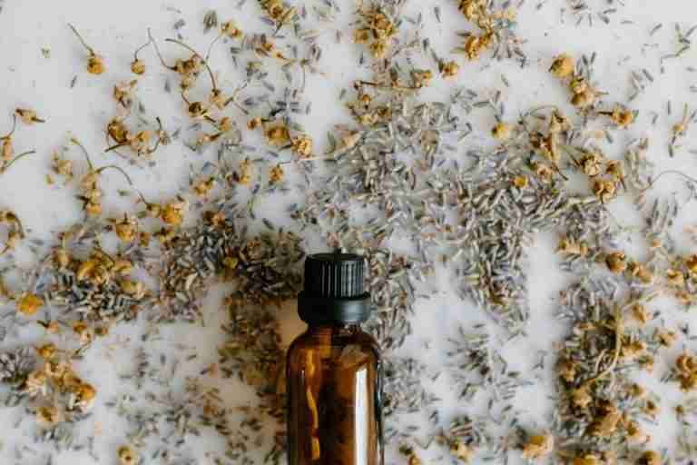 A close-up of an essential oil bottle surrounded by dried lavender and chamomile on a white surface.