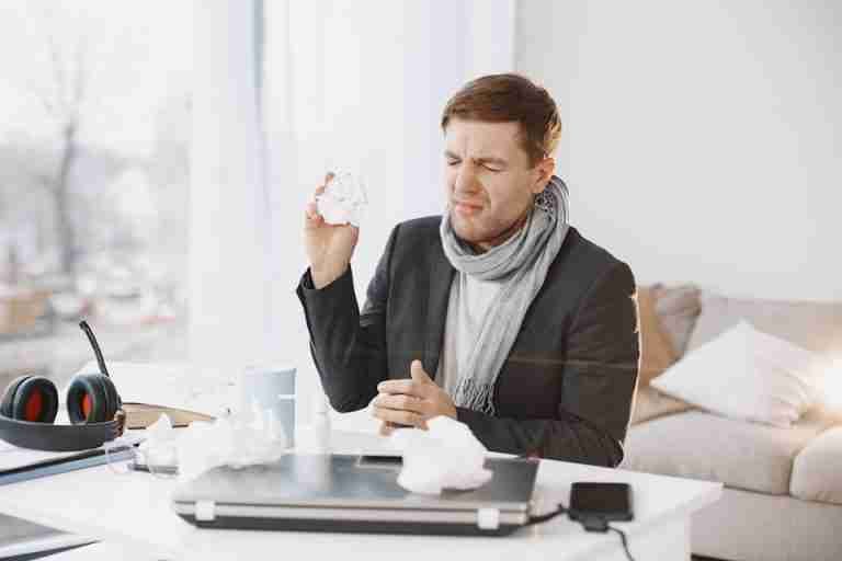 A sick man wearing a scarf sitting at his desk, holding a tissue, displaying signs of discomfort.