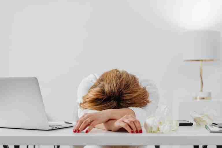 Tired woman resting head on arms at desk with laptop, showing fatigue in modern office.