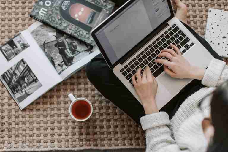 Overhead view of a woman typing on a laptop at home, surrounded by magazines and a cup of tea on the floor.