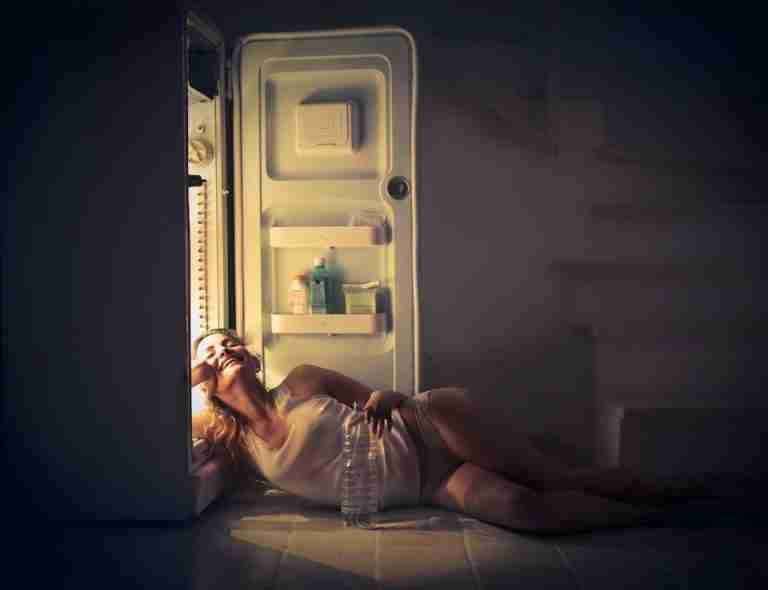 Woman in sleepwear resting next to an open fridge, with a serene smile and a bottle of water.