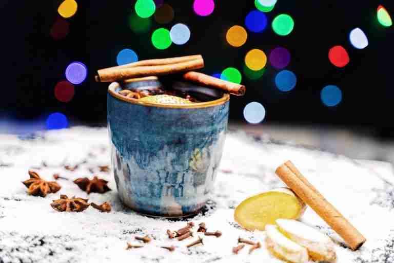 Close-up of a ceramic mug with spiced tea, cinnamon sticks, and ginger slices with bokeh lights in the background.