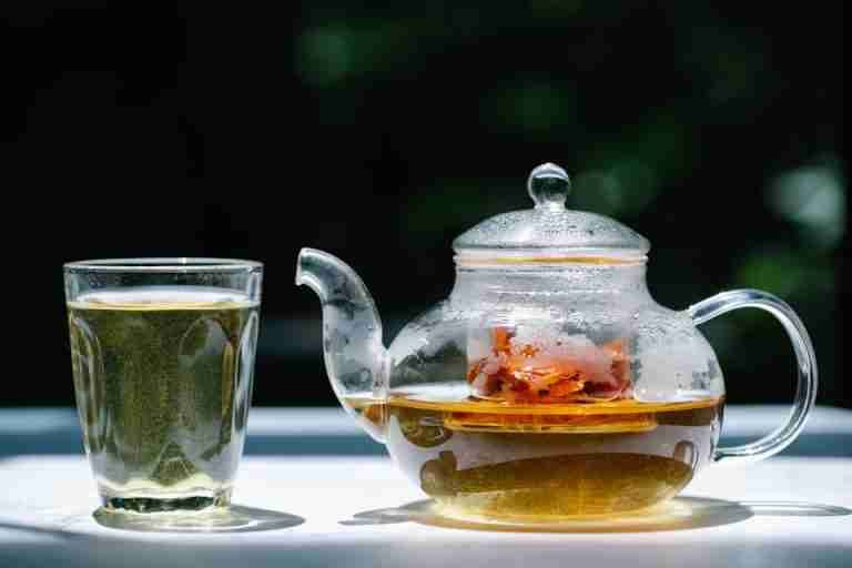 A glass teapot filled with herbal tea next to a transparent glass under sunlight. Refreshing and aromatic.
