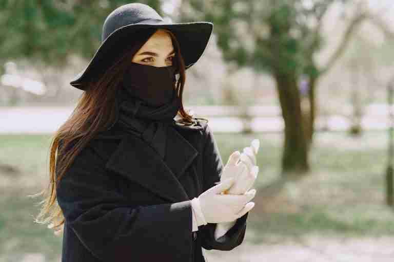 Female in stylish outerwear and hat putting on protective gloves and looking away while walking in park during coronavirus outbreak