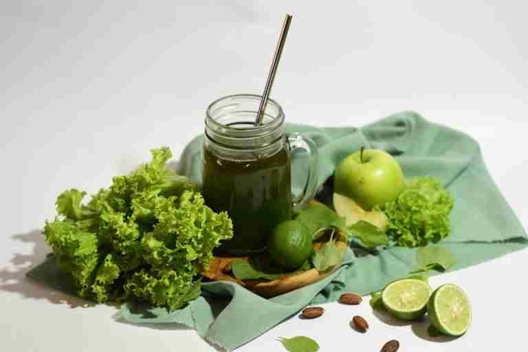 A vibrant green juice surrounded by fresh apples, limes, and lettuce, served in a mason jar with a metal straw.