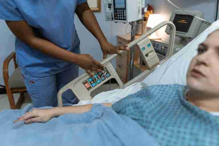 A nurse in scrubs assists a patient in a hospital bed with medical equipment nearby.