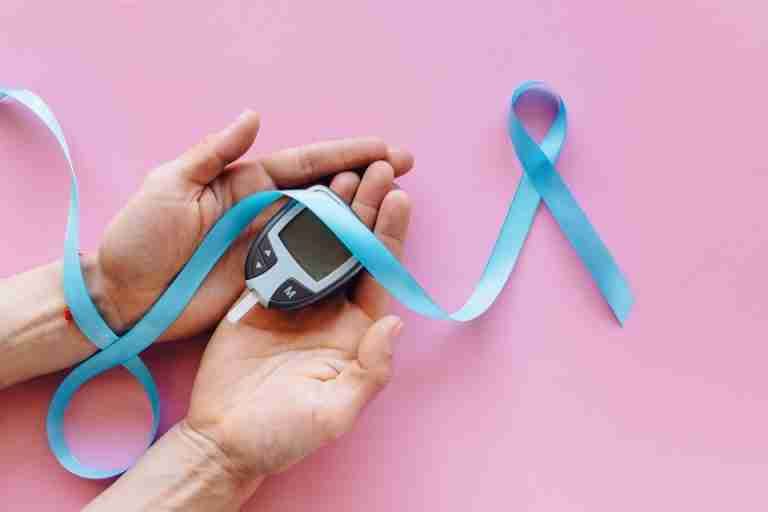 A close-up image of hands holding a glucometer with a blue awareness ribbon on a pink surface, symbolizing diabetes awareness.