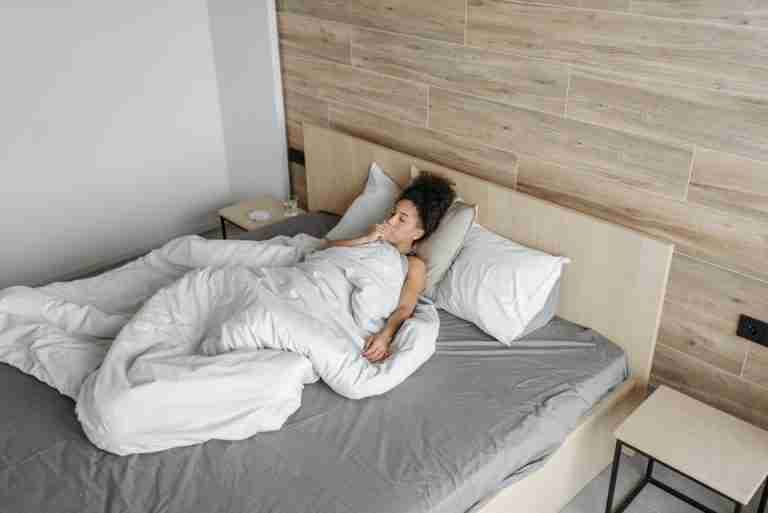A woman lying in bed under a blanket, appearing unwell, in a modern bedroom setting.