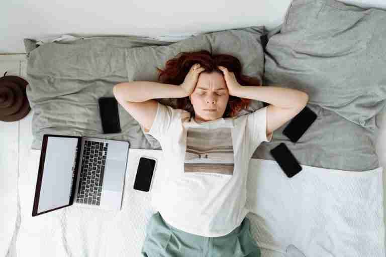 A distressed woman lying in bed surrounded by digital devices, showing signs of stress.