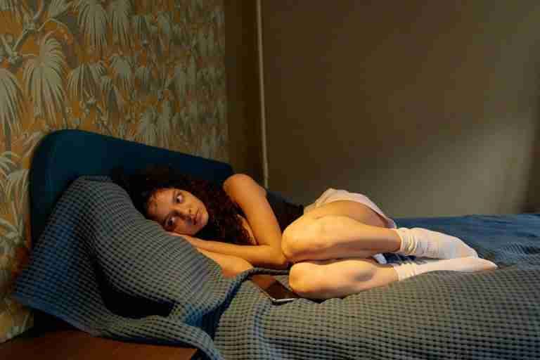 A contemplative young woman lies curled up on a bed indoors, expressing solitude and introspection.