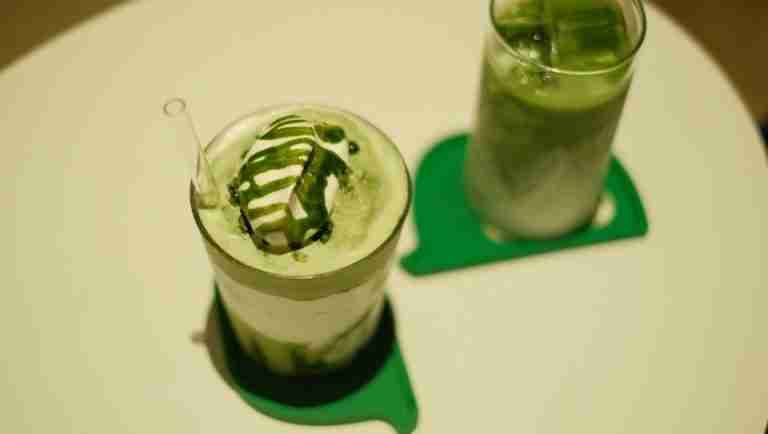 Two iced matcha lattes topped with cream and matcha drizzle on a table.