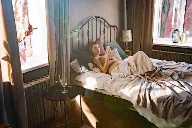 Pregnant woman lying in a cozy bedroom using a cellphone, enjoying a tranquil morning light.