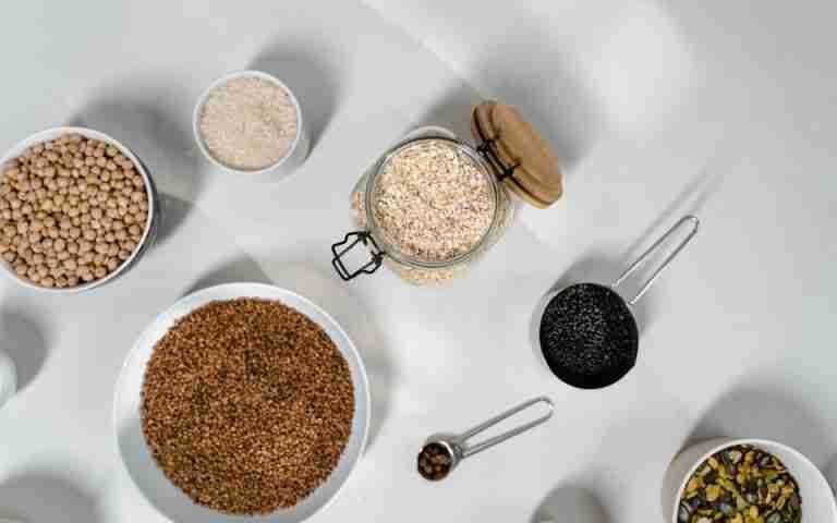 Top view of various grains and seeds in bowls and jars on a white surface.