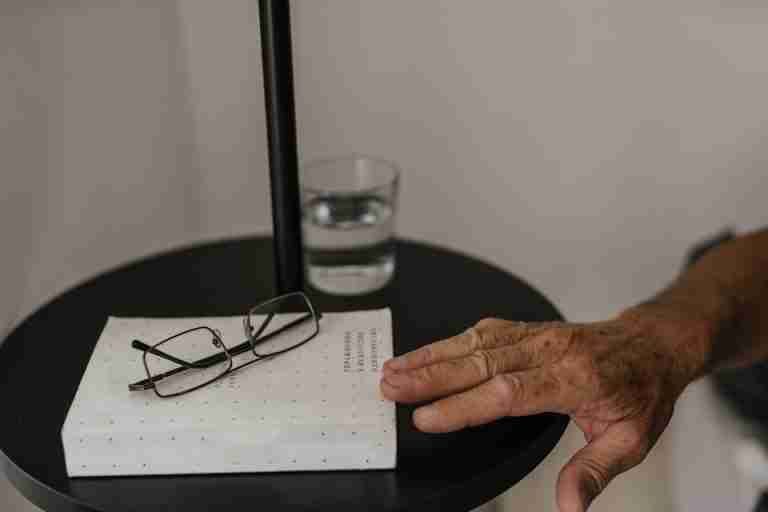 Close-up of an elderly hand reaching for glasses on a bedside table with a book and water glass, in a calm setting.