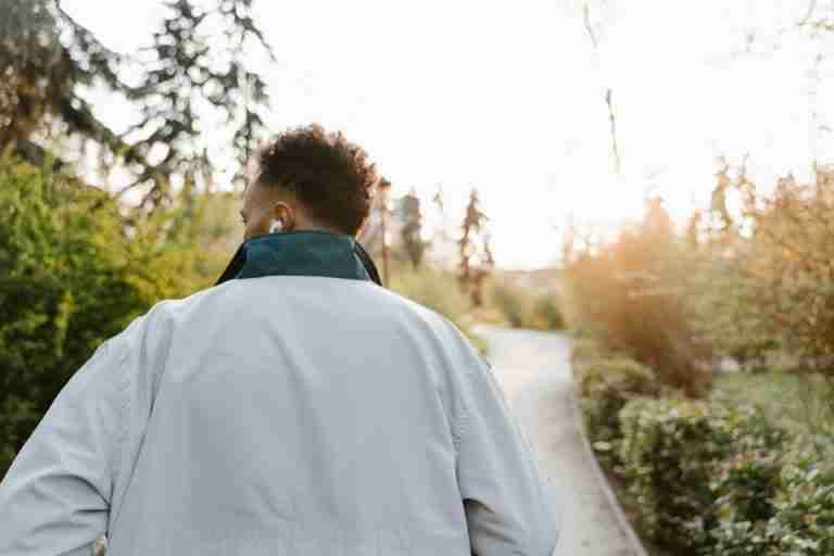 A man with headphones walking in a sunlit park, wearing a white jacket.
