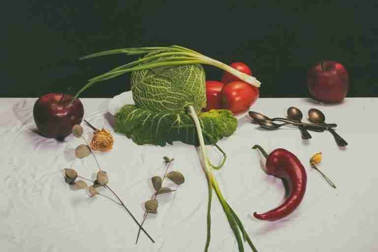 A vibrant still life of vegetables and fruits on a white tablecloth, featuring cabbage and apples.