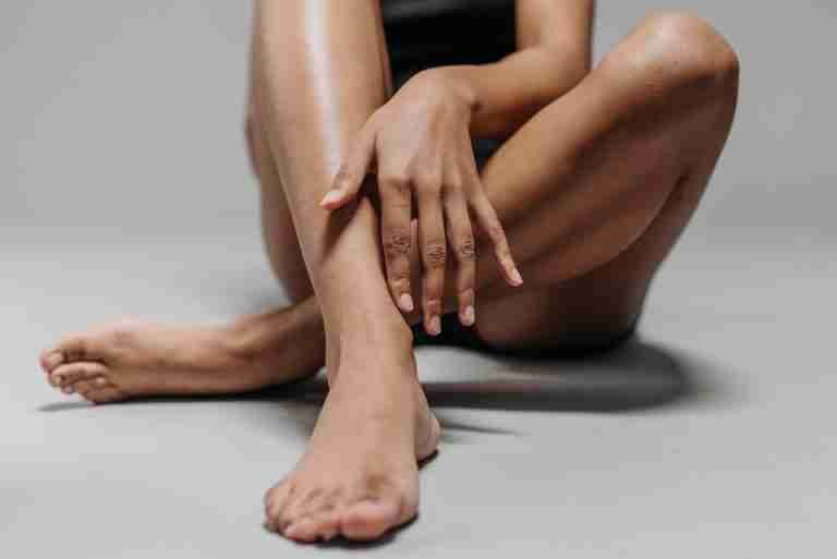 Close-up view of a woman's legs and hands sitting cross-legged on the floor, emphasizing skin tone and texture.