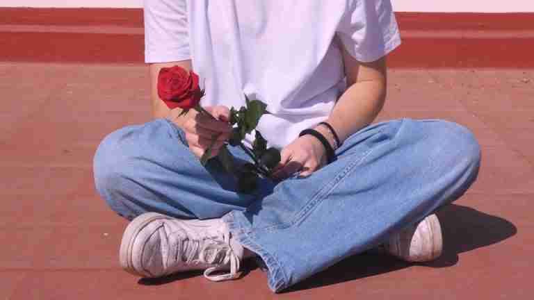 A person in denim and white shirt holding a red rose while sitting cross-legged outdoors.