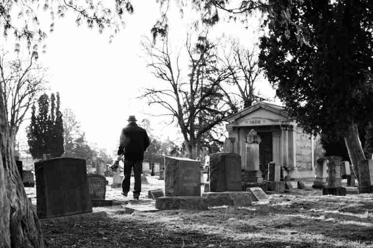 A lone figure walks through a historic cemetery, surrounded by tombstones in a black and white setting.