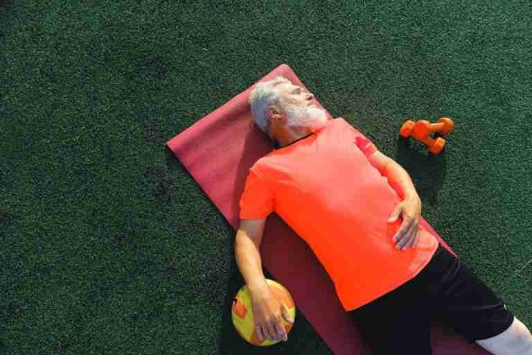 From above of tired senior sportsman lying on fitness mat near ball and dumbbells and resting after training in sunny day