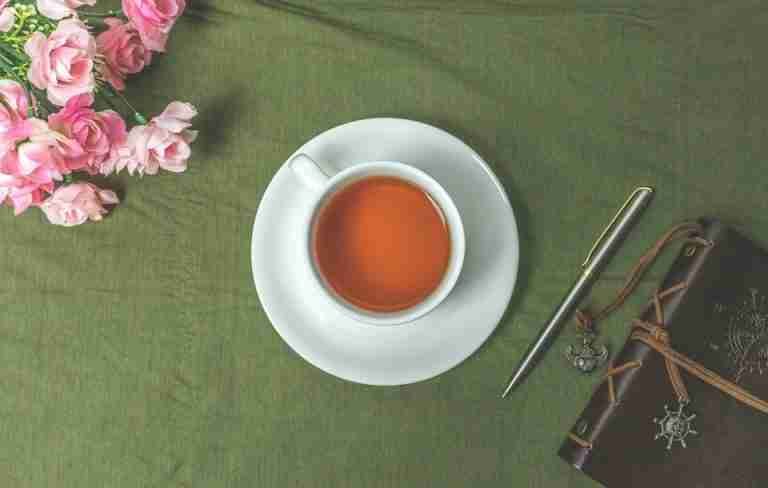 Aesthetic flat lay featuring a cup of tea, pink flowers, pen, and journal on a green background.