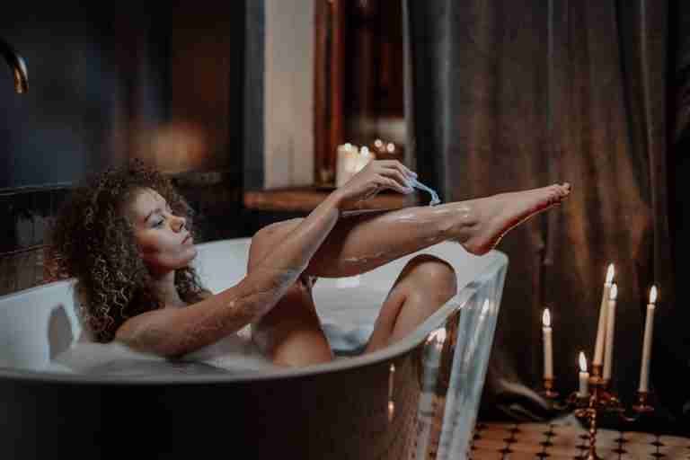 A serene scene of a woman relaxing in a bathtub with candlelight, focusing on shaving.