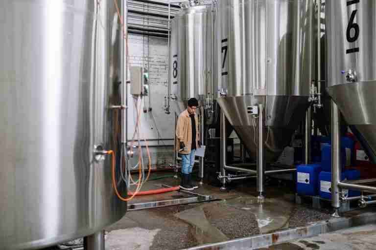 An industrial worker inspects fermentation tanks in a brewery, surrounded by equipment and machinery.