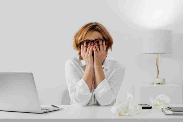 Woman with hands on face, sitting at desk with laptop and tissues, feeling stressed.