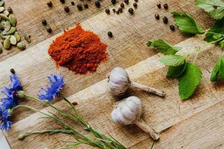 Fresh garlic, paprika, lemon balm, and cornflowers arranged with peppercorns on a rustic wooden board.