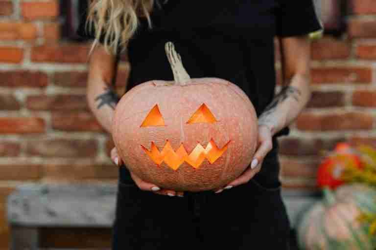 A woman with tattoos holds a glowing jack-o'-lantern in a rustic outdoor setting, perfect for Halloween-themed imagery.