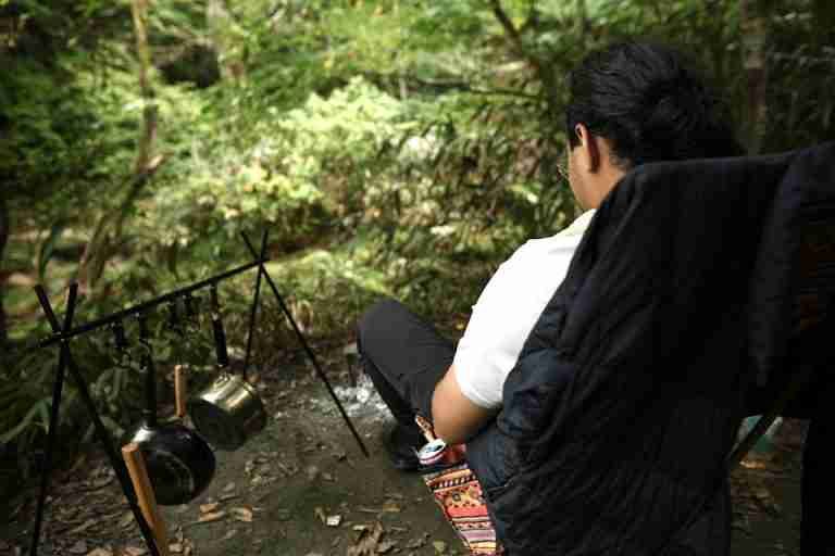 A person relaxes at a campsite with hanging pots and forest surroundings.