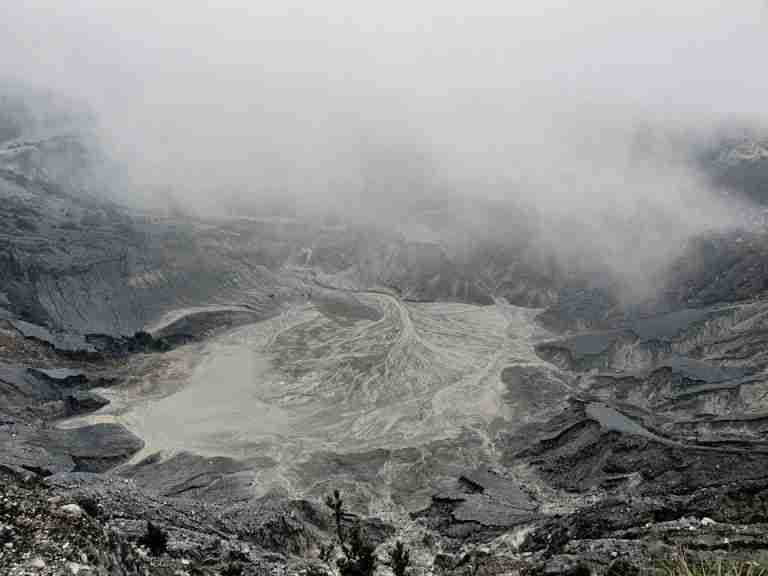From above severe view of rough volcanic crater with hot foamy water and rocky peaks covered with dense mysterious haze