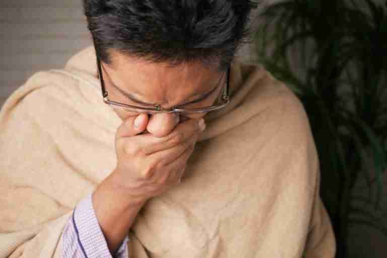 Adult wearing glasses coughs into hand indoors, indicating illness or flu symptoms.