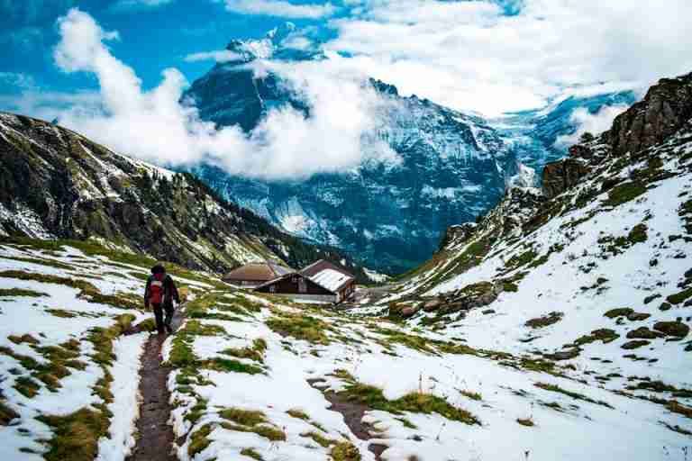 A lone hiker traverses a scenic mountain path with snow and clouds around.