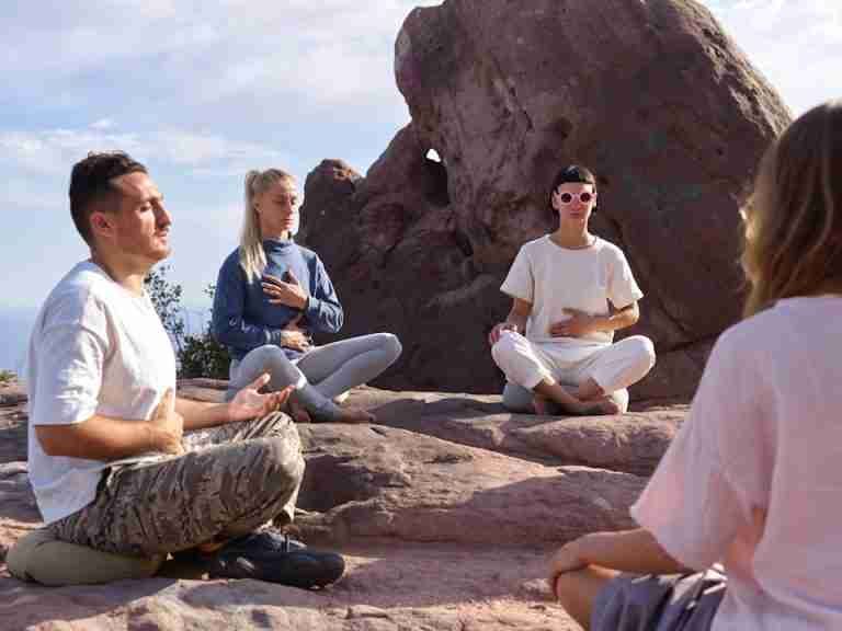 Four people meditate on rocky terrain under a clear sky, promoting serenity and mindfulness.