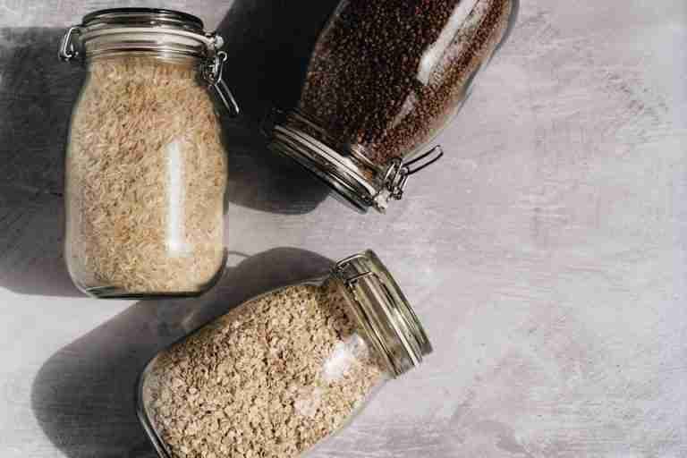 Top-down view of rice, oats, and buckwheat in glass jars on a textured surface.