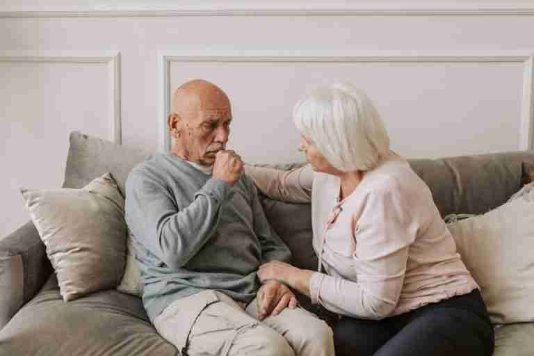 An elderly woman comforts a man coughing on a couch, showcasing care and affection.