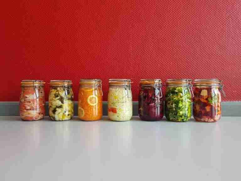 A row of vibrant, multicolored mason jars filled with assorted fermented vegetables against a red wall.