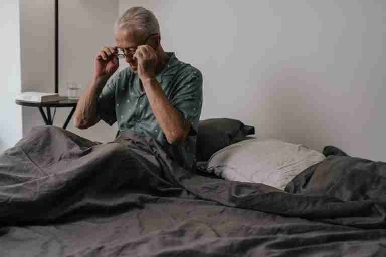 Senior man adjusts glasses while sitting in bed, expressing morning routine and daily life.