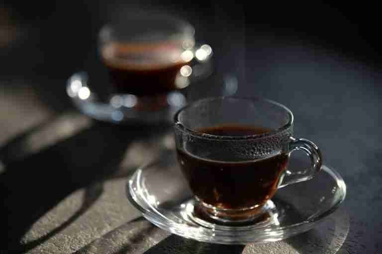 Close-up of steaming black coffee in elegant glass cup with saucer, captured in soft light.