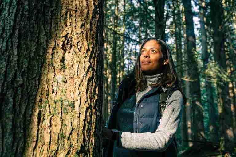 A woman standing by a tree, enjoying nature in a serene forest setting.